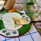 A wheel of soft white cheese with a slice cut out, served on a decorative green and white plate with a small gold cheese knife, set on a patterned tablecloth.