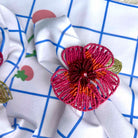 A close-up of a decorative beaded flower napkin ring in shades of magenta, orange, and gold, resting on a white napkin with a blue grid and strawberry print.