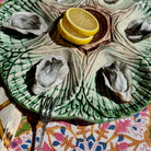 A green and beige ceramic oyster shell platter with a leaf-like embossed pattern, served with four raw oysters and two lemon slices on a patterned tablecloth.