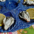 A close-up of a blue ceramic platter holding two raw oysters on the half, garnished with lemon slices, with a small bowl and spoon at the top.