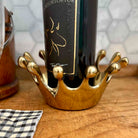 A gold crown-shaped wine bottle holder cradling a dark wine bottle on a wooden surface with a tiled background.