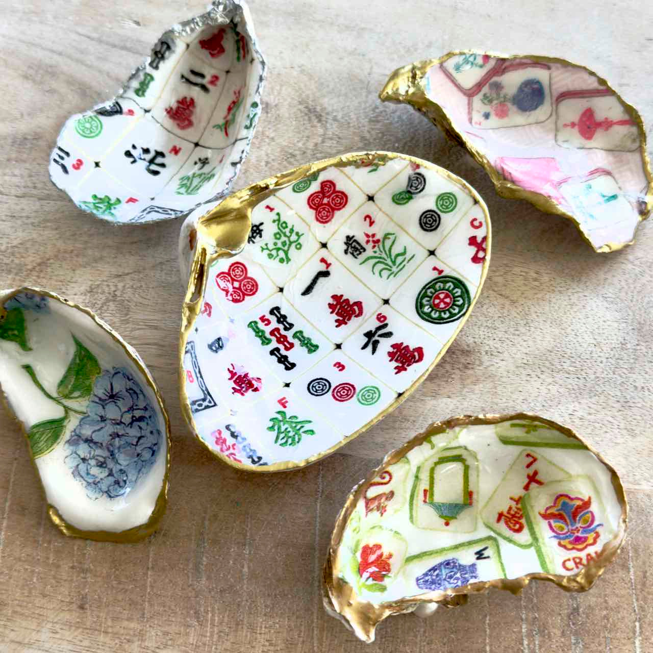 Five small decorative shell-shaped dishes with gold-edged interiors, decorated with colorful printed patterns including mahjong tiles, floral motifs, and playing cards, arranged on a light wood surface.