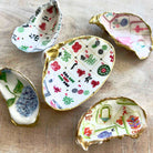 Five small decorative shell-shaped dishes with gold-edged interiors, decorated with colorful printed patterns including mahjong tiles, floral motifs, and playing cards, arranged on a light wood surface.