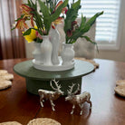 Decorative table setting with white vases, flowers, and deer figurines on a wooden surface.