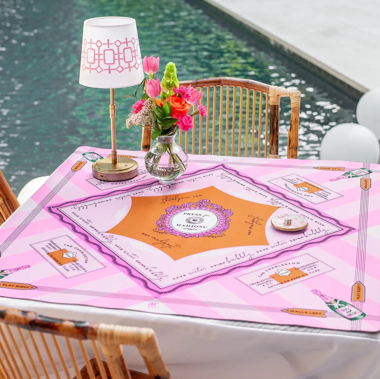 Table with a pink tablecloth featuring a decorative design, lamp, and flowers by a pool.