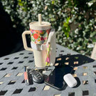 Floral-patterned tumbler with a straw, phone, keys, and lip gloss on a table outdoors.