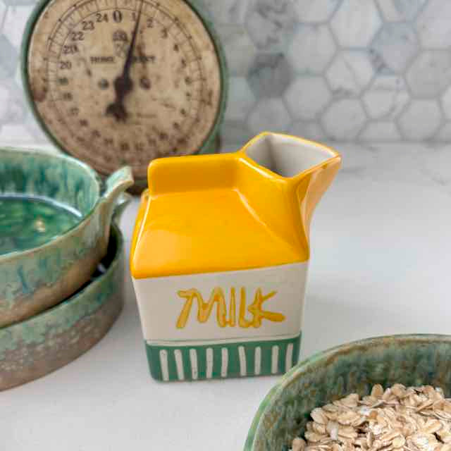 Ceramic milk carton with 'Milk' written on it, placed on a surface with bowls and a scale in the background.