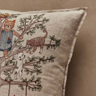 A close-up of a beige linen throw pillow featuring embroidered cats and a dog climbing a tree, set against a matching beige background.