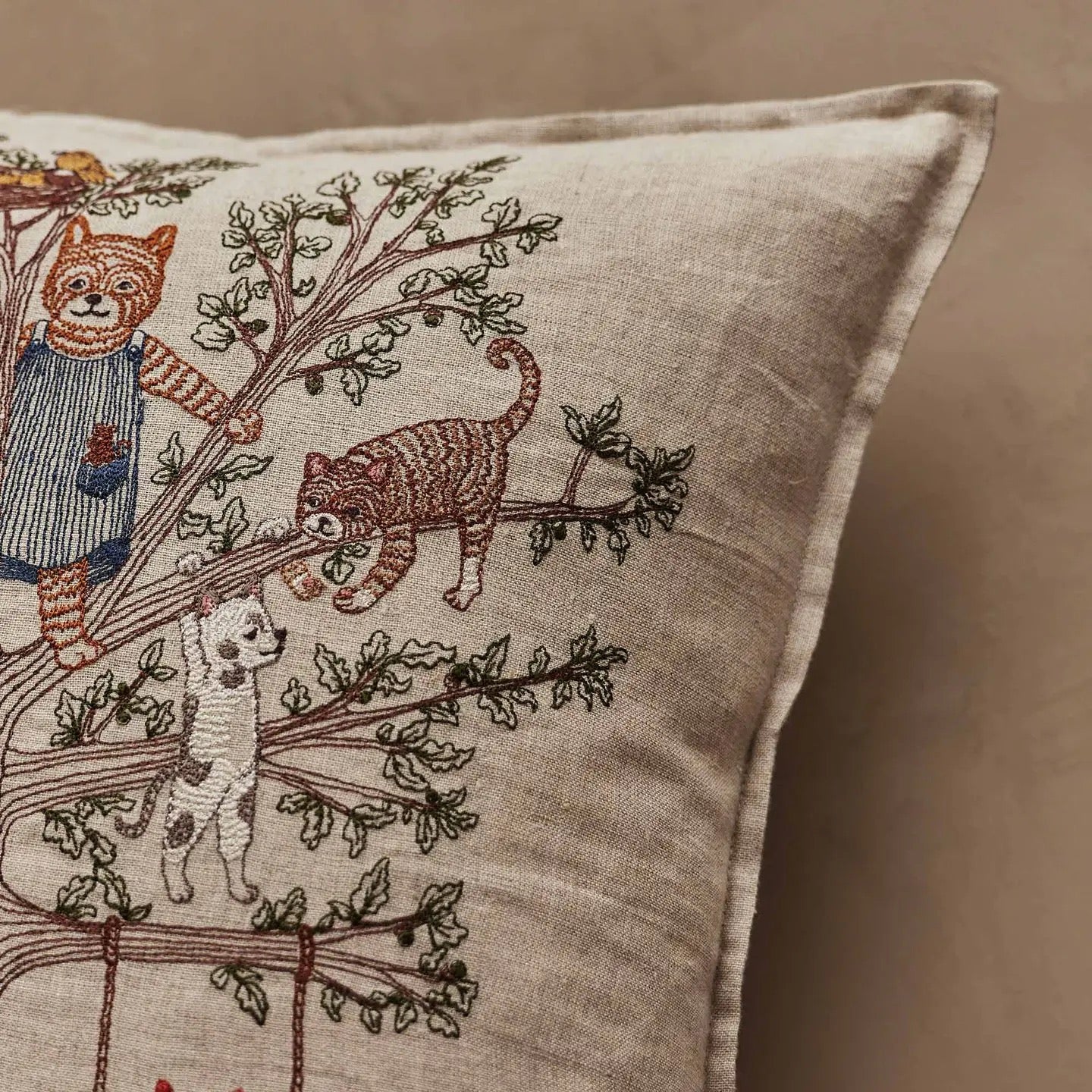 A close-up of a beige linen throw pillow featuring embroidered cats and a dog climbing a tree, set against a matching beige background.