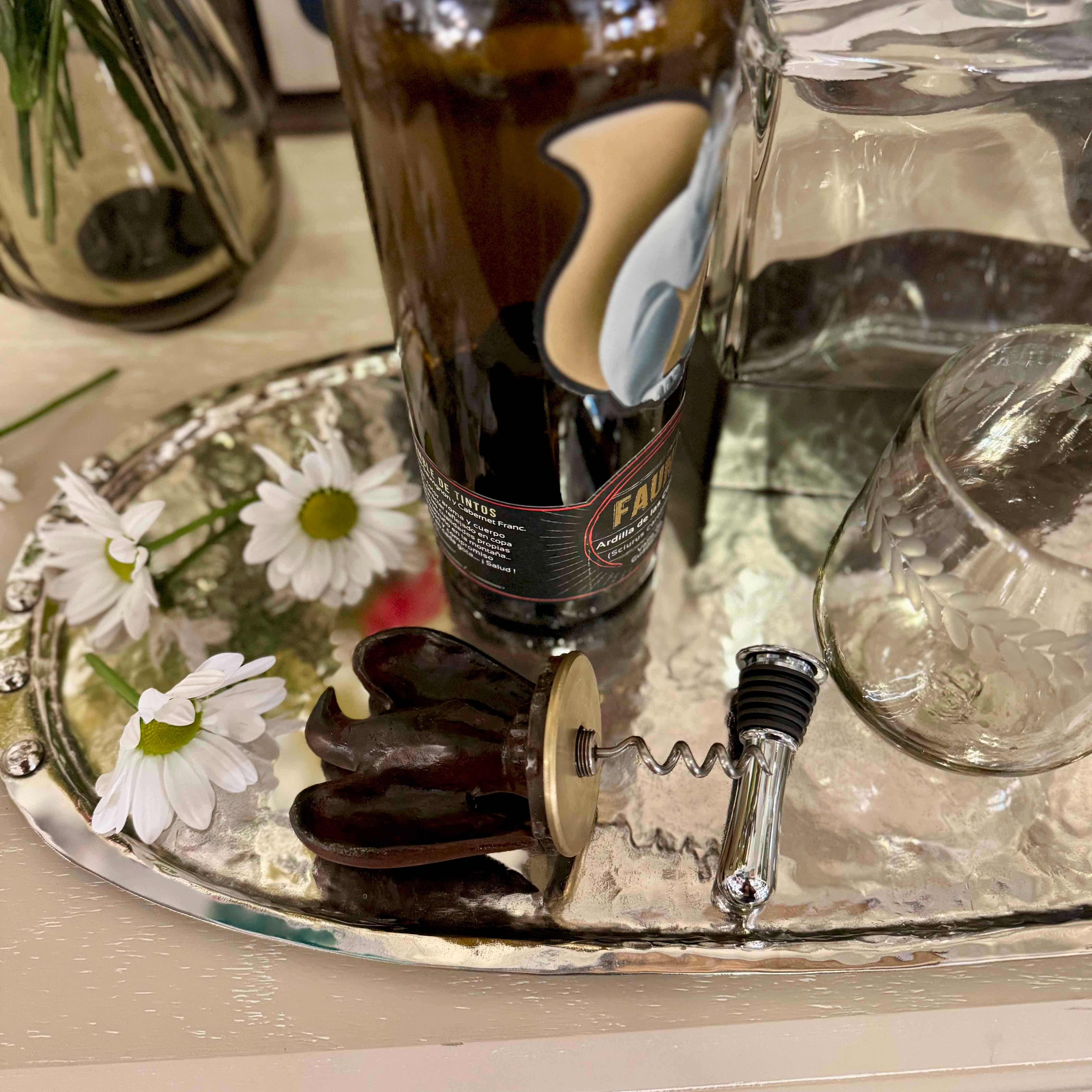 Bottle of wine, corkscrew, and daisies on a silver tray with a blurred background.