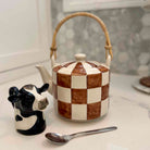Checkered teapot with a cow-shaped salt shaker and spoon on a marble surface.