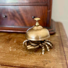 A brass crab-shaped bell holder with a dome-shaped top and metallic claws, resting on a worn wooden surface with a blurred wooden cabinet in the background.