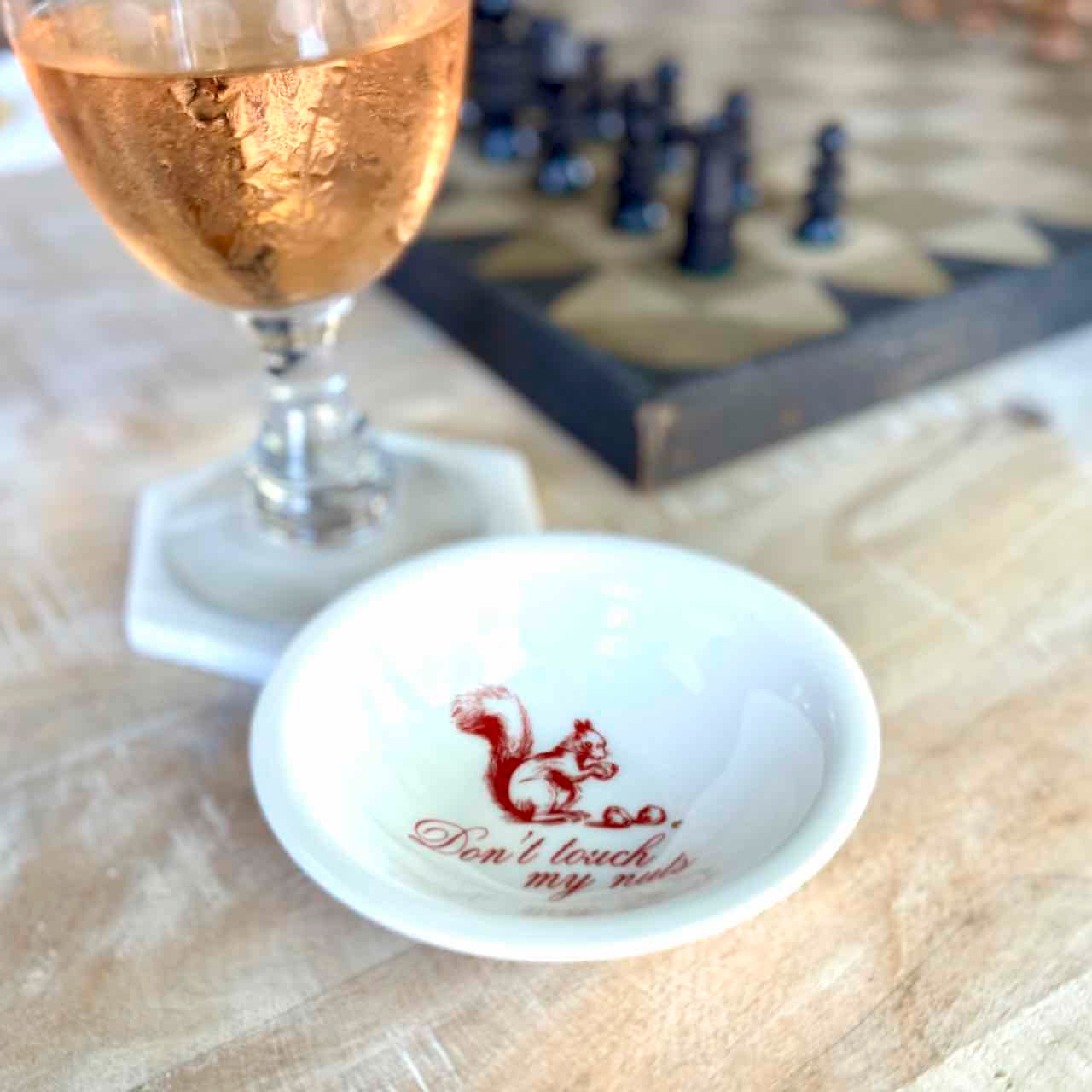 A small white ceramic bowl with a red printed squirrel and the phrase 'Don't touch my nuts' sits on a wooden surface next to a glass of rosé wine, with a blurred chess game in the background.