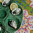 Three raw oysters served on a green leaf-shaped ceramic platter with lemon slices, set on a patterned tablecloth.