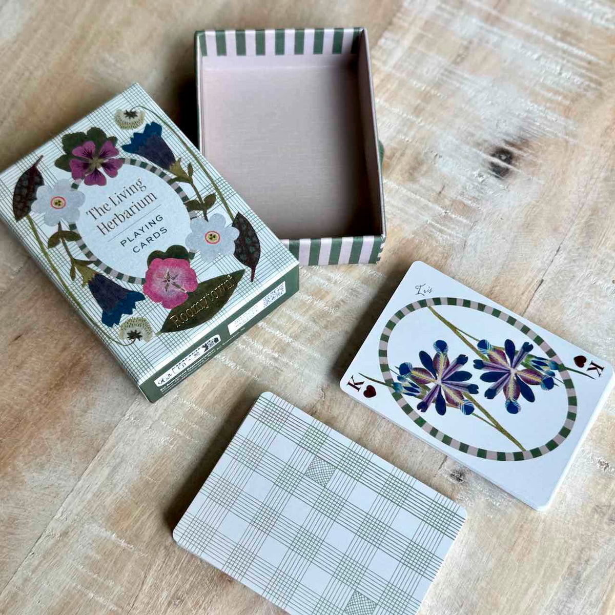 The Living Herbarium Playing Cards set displayed on a light wood surface, showing the illustrated outer box, an open striped interior box, a card face with floral artwork, and a card back with a plaid pattern.