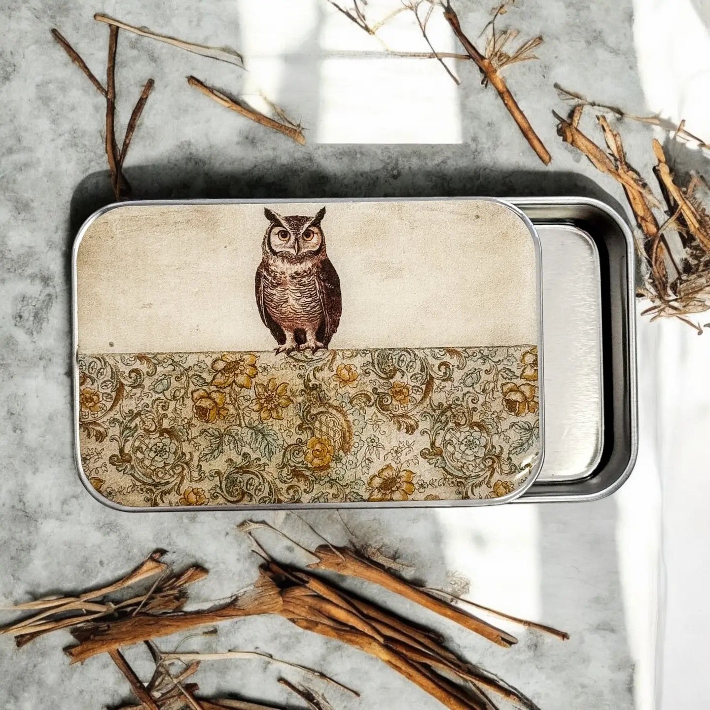 Metal tin with owl design on a textured surface with twigs