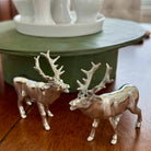 Two silver deer figurines on a wooden surface with a white decorative object in the background.