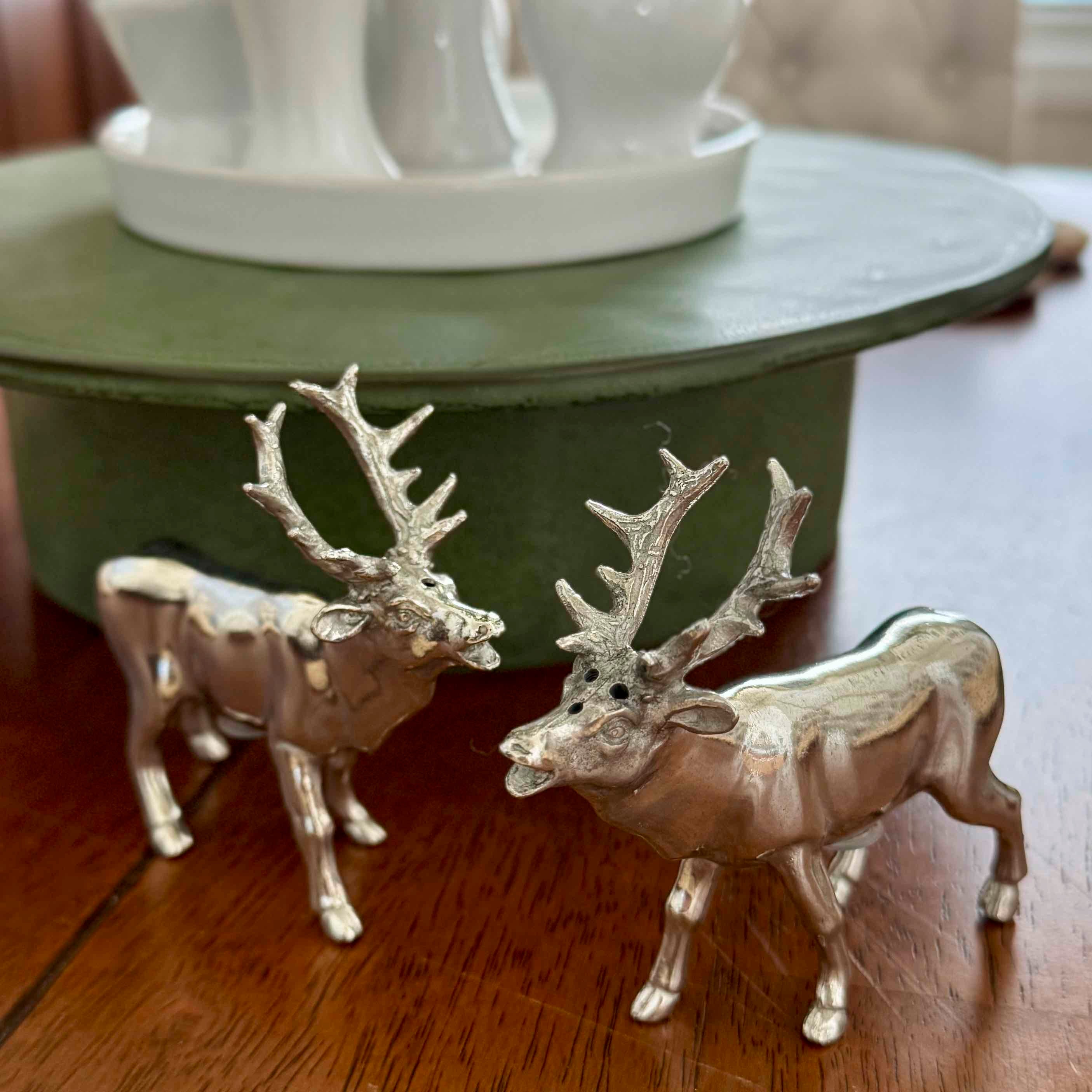 Two silver deer figurines on a wooden surface with a white decorative object in the background.