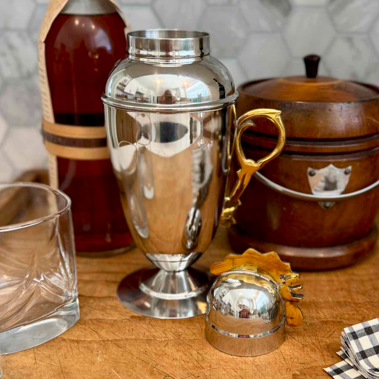 A polished silver metal cocktail shaker with a gold-tone handle and matching small metal scoop, displayed on a wooden surface with a glass, amber bottle, and wooden container in the background.