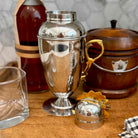 A polished silver metal cocktail shaker with a gold-tone handle and matching small metal scoop, displayed on a wooden surface with a glass, amber bottle, and wooden container in the background.