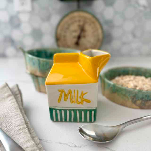 Ceramic milk carton with 'Milk' text on a kitchen counter with bowls and a spoon.