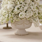 A white ceramic pedestal planter with ornate embossed detailing, filled with dense white faux flowers, set on a light beige surface.