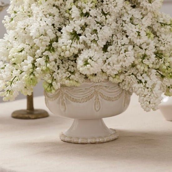 A white ceramic pedestal planter with ornate embossed detailing, filled with dense white faux flowers, set on a light beige surface.
