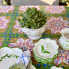 A collection of white ceramic tableware with green botanical illustrations, including a small flower pot with a succulent, a stack of matching saucers, and a creamer pitcher, arranged on a colorful floral tablecloth.