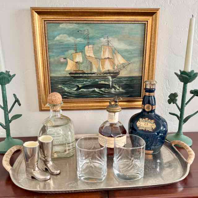 Tray with bottles and glasses in front of a framed painting of a ship.