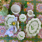 A coordinated set of white ceramic tableware with green botanical illustrations, arranged on a colorful floral tablecloth. The set includes platters, bowls, a small pitcher, and a napkin with a flower centerpiece.