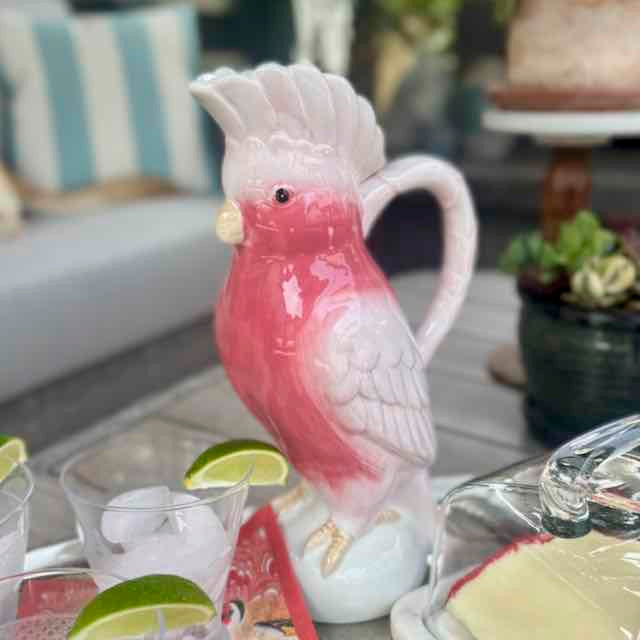 Pink and white ceramic parrot-shaped teapot on a table with drinks and snacks.