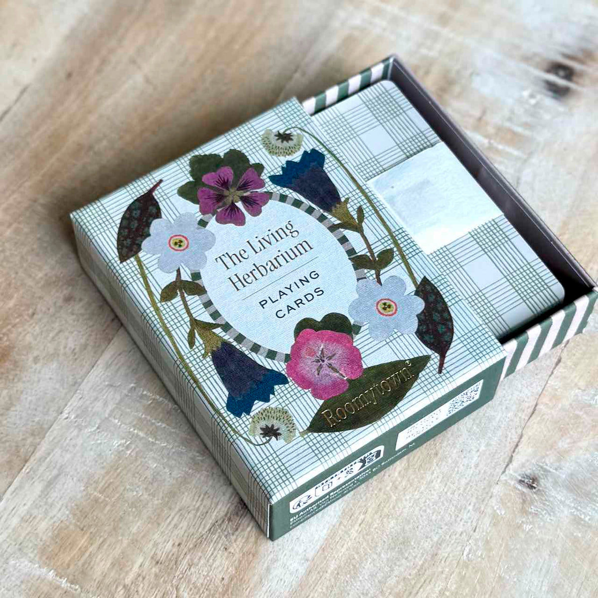 The Living Herbarium Playing Cards deck in its illustrated box, photographed on a light wood tabletop. The box features a floral and botanical design with plaid accents and the title 'The Living Herbarium Playing Cards' visible on the front.