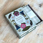 The Living Herbarium Playing Cards deck in its illustrated box, photographed on a light wood tabletop. The box features a floral and botanical design with plaid accents and the title 'The Living Herbarium Playing Cards' visible on the front.