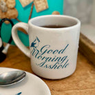 A white ceramic coffee mug with a dark blue graphic of a bird and the text 'Good Morning Asshole' printed on it, filled with coffee and resting on a wooden surface.