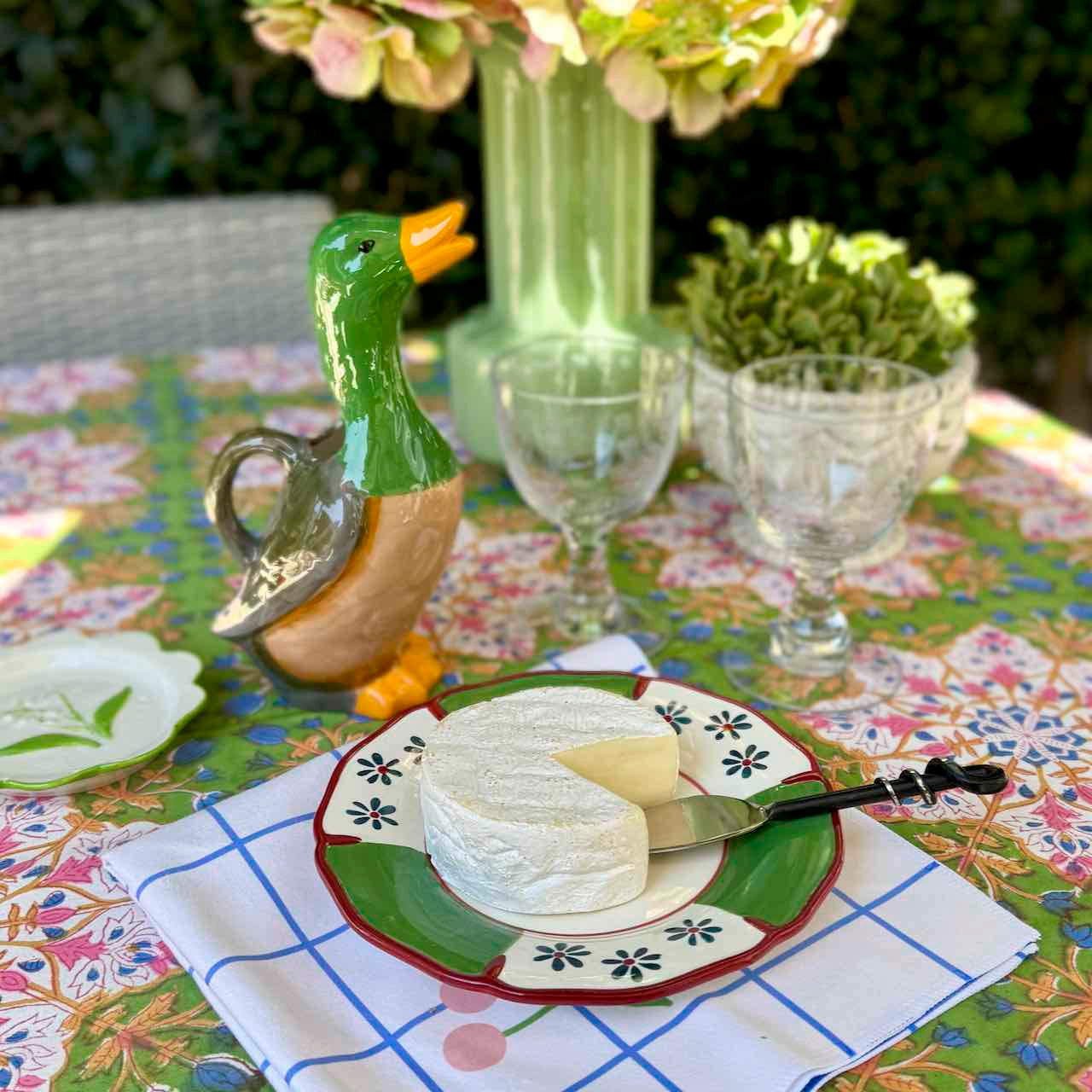 A styled outdoor dining table setting featuring a ceramic duck-shaped pitcher, a wheel of soft cheese on a green and red patterned plate, two clear glass goblets, and a colorful floral tablecloth.