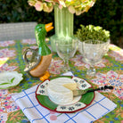 A styled outdoor dining table setting featuring a ceramic duck-shaped pitcher, a wheel of soft cheese on a green and red patterned plate, two clear glass goblets, and a colorful floral tablecloth.