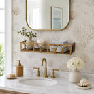 A styled bathroom vanity featuring a white marble countertop with a brass faucet, a woven soap dispenser, a round mirror with a gold frame, and a bamboo wall shelf holding folded towels and a candle. The wall is covered in beige wallpaper with a botanical shell and coral pattern.