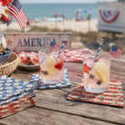 Two glasses of lemonade with fruit and American flags on a wooden table by the beach.