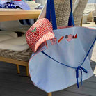 A blue and white striped tote bag with blue straps and embroidered summer-themed patches, shown on a patio next to a red-and-white gingham sun hat.