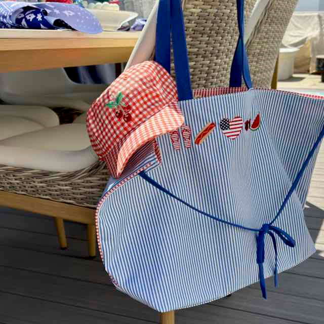 A blue and white striped tote bag with blue straps and embroidered summer-themed patches, shown on a patio next to a red-and-white gingham sun hat.