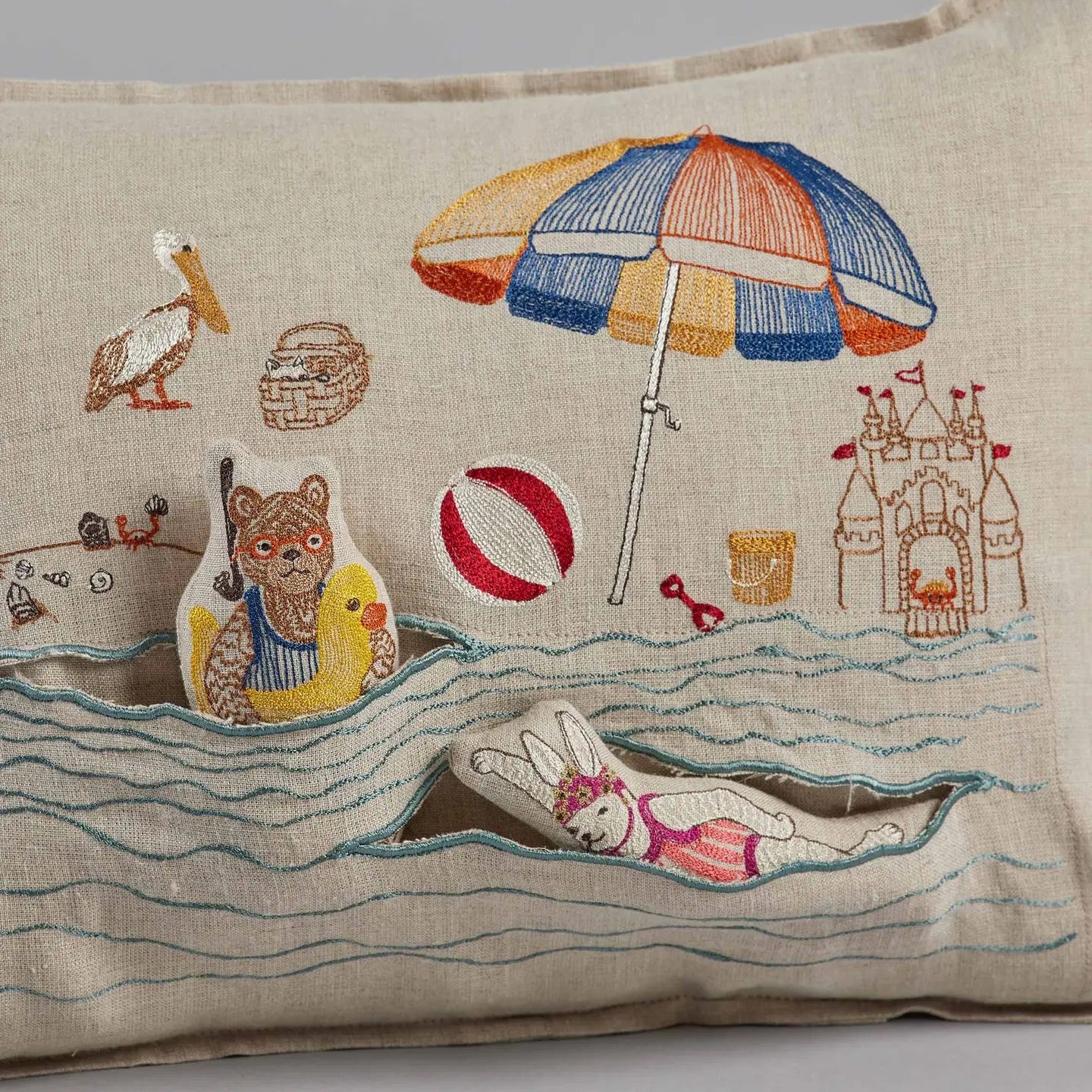 Embroidered pillow with beach-themed design featuring animals, an umbrella, and sandcastles.