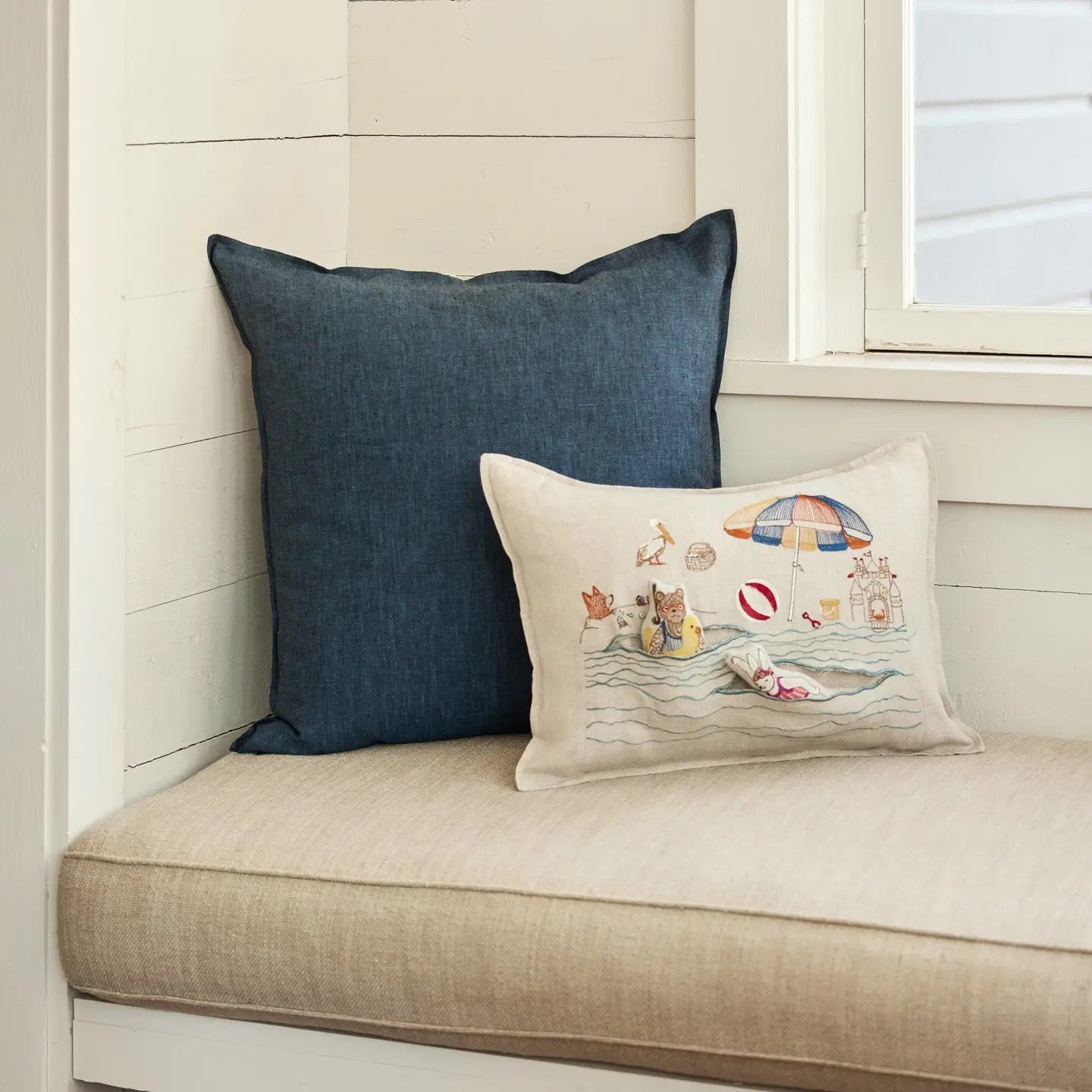 Two pillows on a cushioned bench with a blue pillow and a decorative pillow with a beach scene.