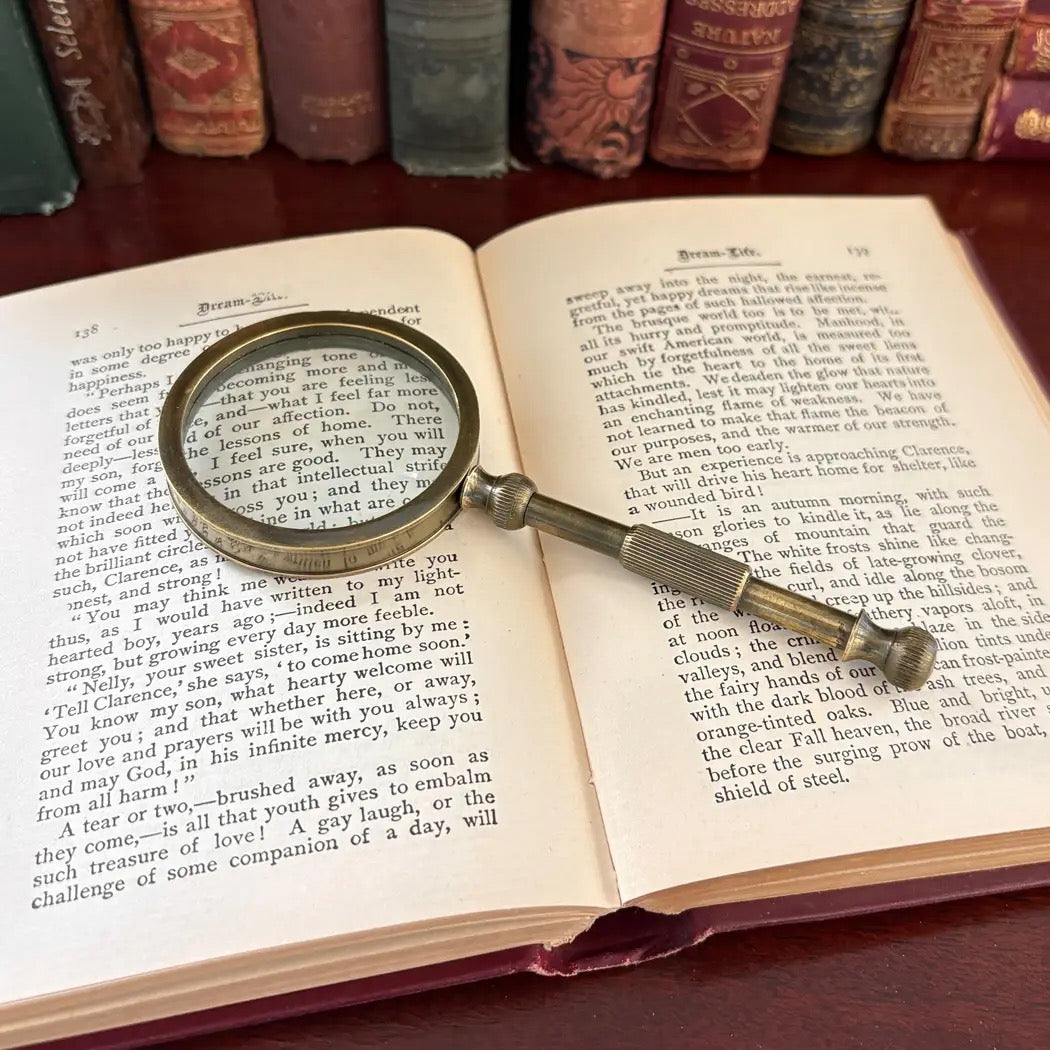 A vintage-style brass magnifying glass with a decorative key-shaped handle rests on the pages of an open book, with a green Bible and a red patterned book in the blurred background.