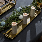 Three lit pillar candles arranged on a long gold tray with evergreen branches and decorative ornaments on a dark tablecloth.