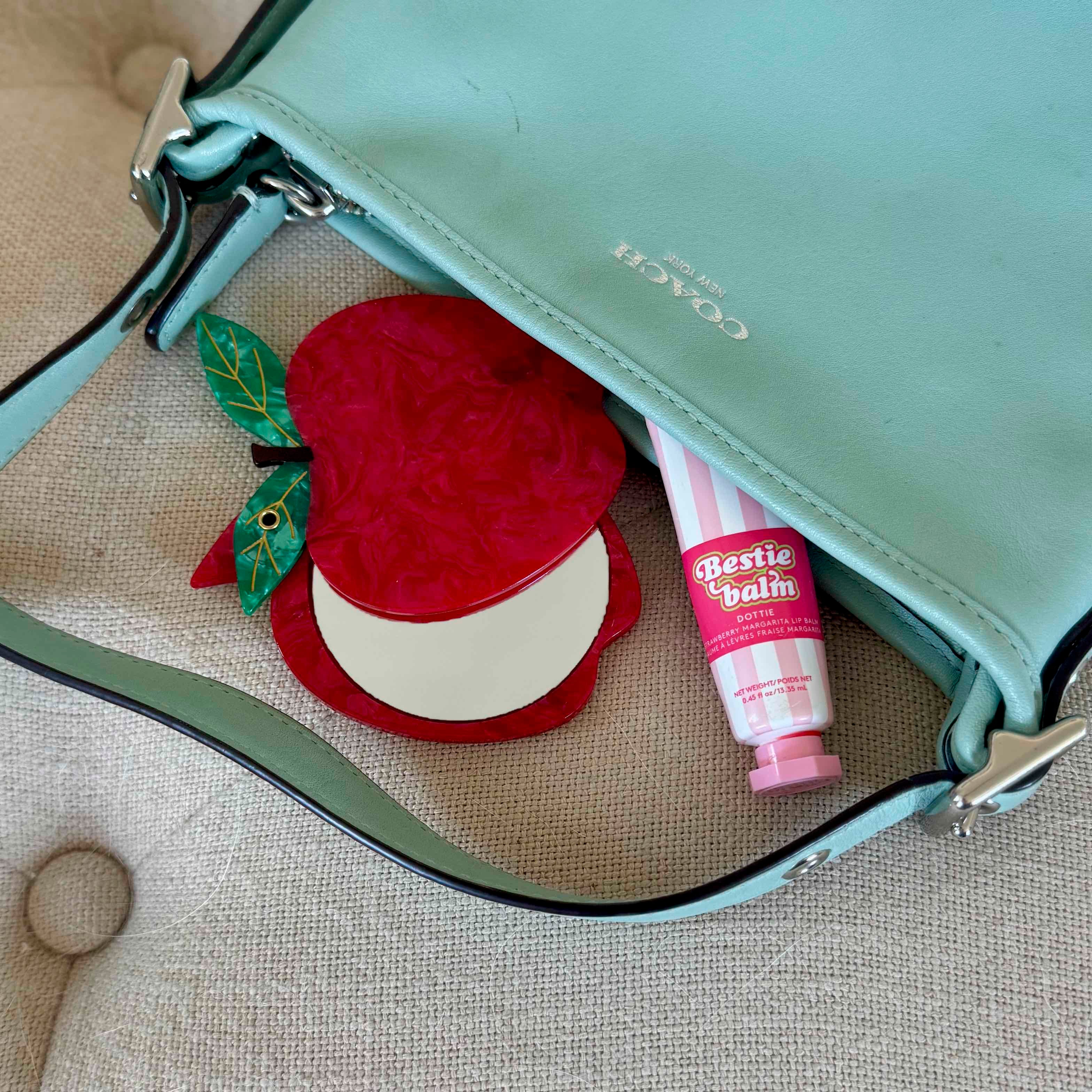 Green handbag with a red apple-shaped item and pink tube of balm on a beige surface