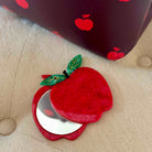 Red apple-shaped compact mirror with a green leaf on a beige surface.
