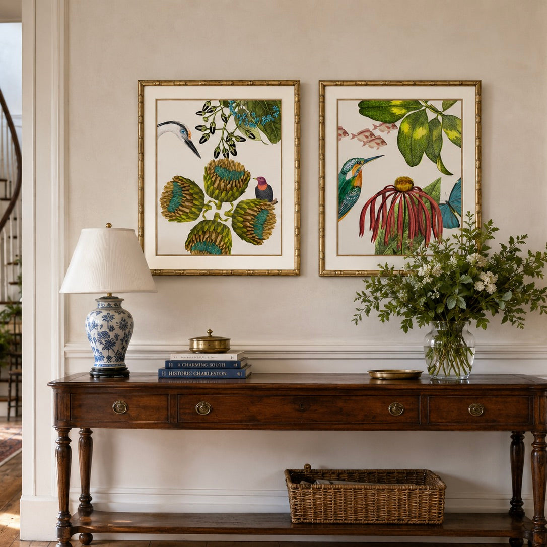 Two framed botanical and bird art prints hang on a beige wall above a dark wood console table. The left print shows a bird and green seed pods, and the right print shows a hummingbird with green leaves and red-pink flowers.