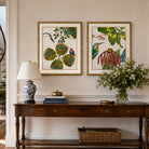Two framed botanical and bird art prints hang on a beige wall above a dark wood console table. The left print shows a bird and green seed pods, and the right print shows a hummingbird with green leaves and red-pink flowers.