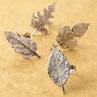 Four silver-toned leaf-shaped earrings arranged on a textured beige background.