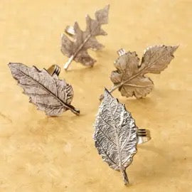 Four silver-toned leaf-shaped earrings arranged on a textured beige background.